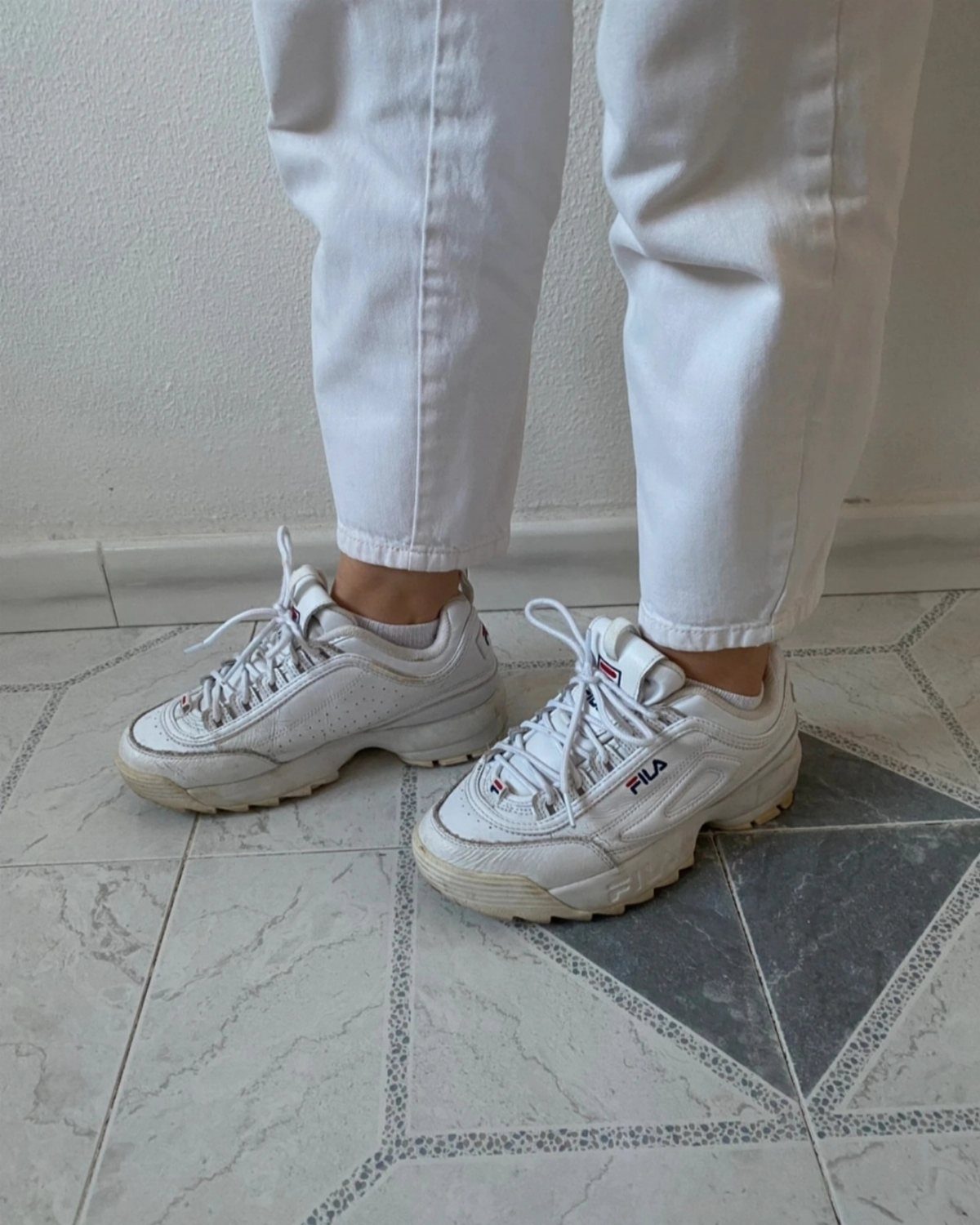 Person wearing white jeans and chunky white Fila sneakers on a tiled floor.