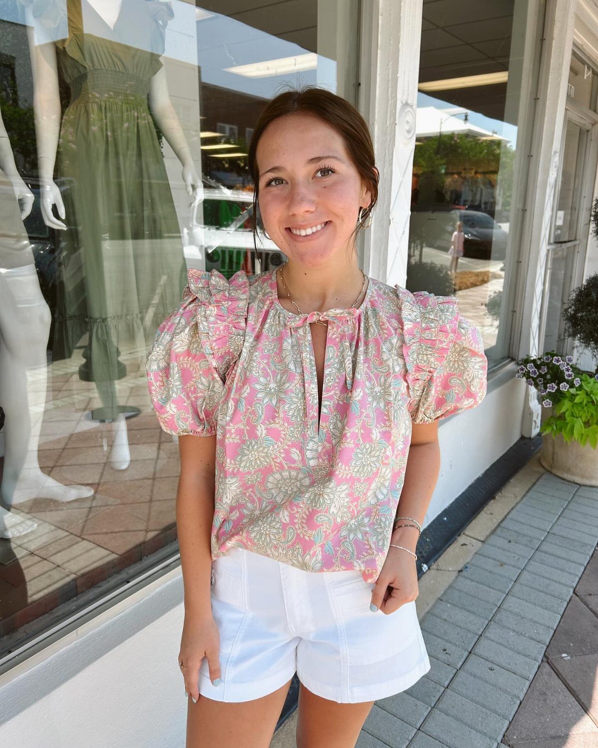 A woman smiling in a pink patterned blouse and white shorts standing outdoors.