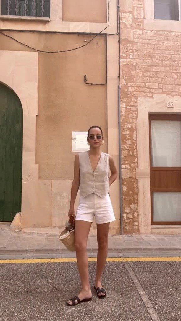 Woman in a cream vest and white shorts standing on a street by stone buildings.