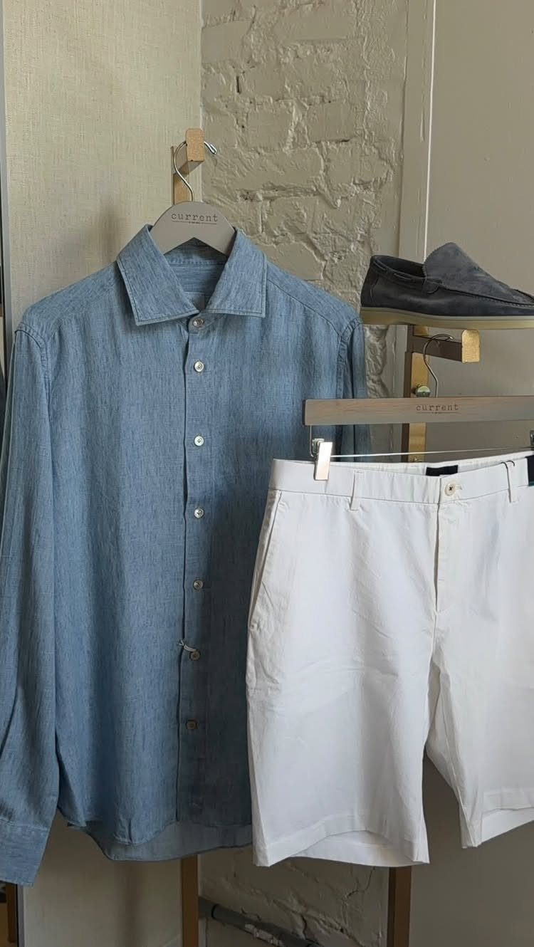 A blue chambray button-down shirt hanging next to white shorts on a clothing rack.