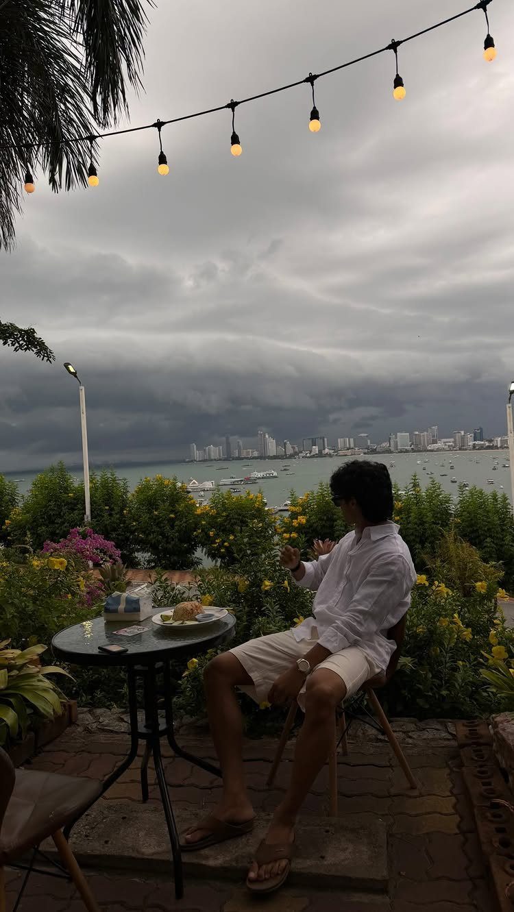 Man in white linen shirt and shorts sitting at an outdoor cafe table by the water.