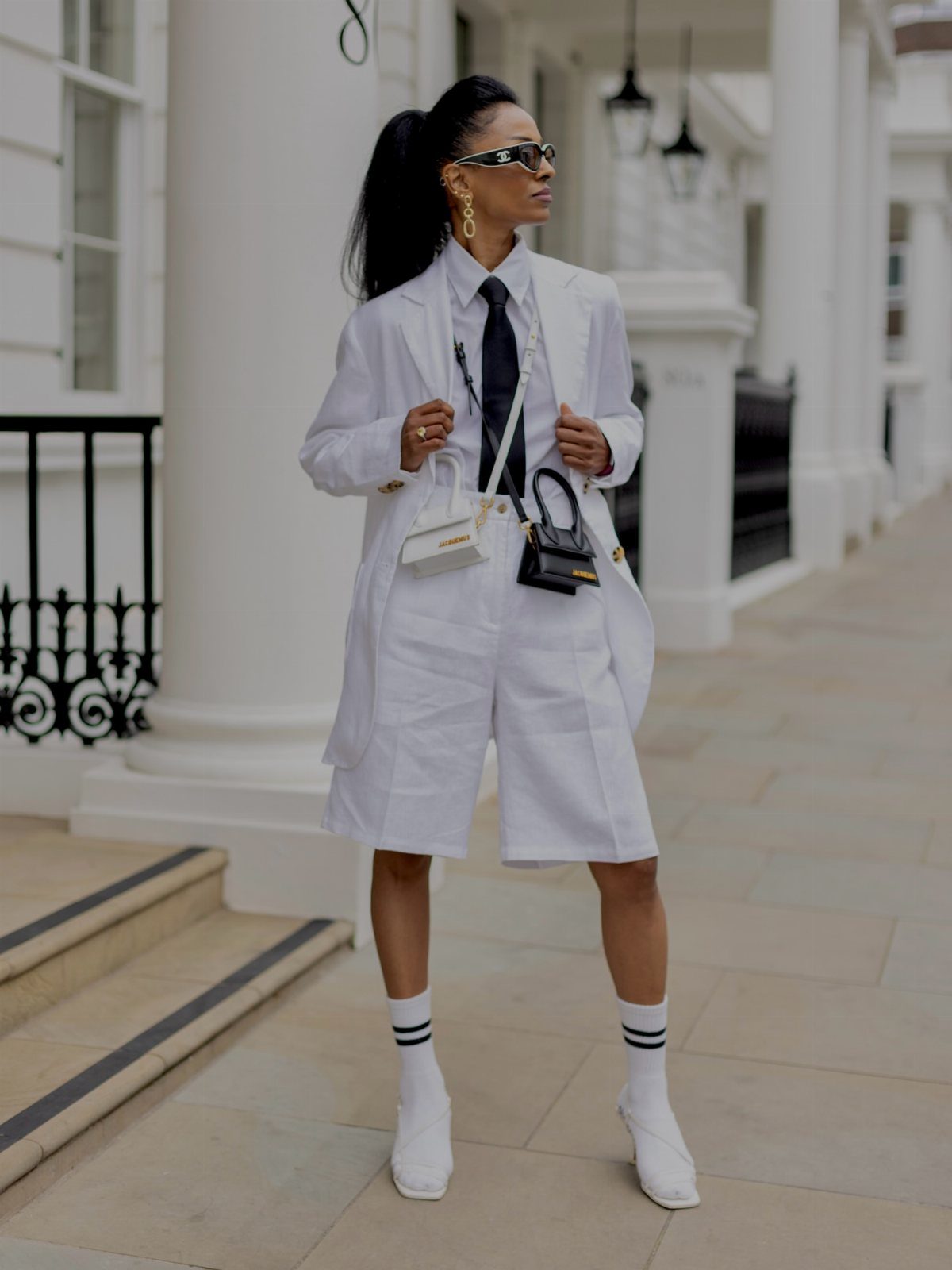 Woman posing in an all-white blazer, shirt, and shorts outfit with a black tie.