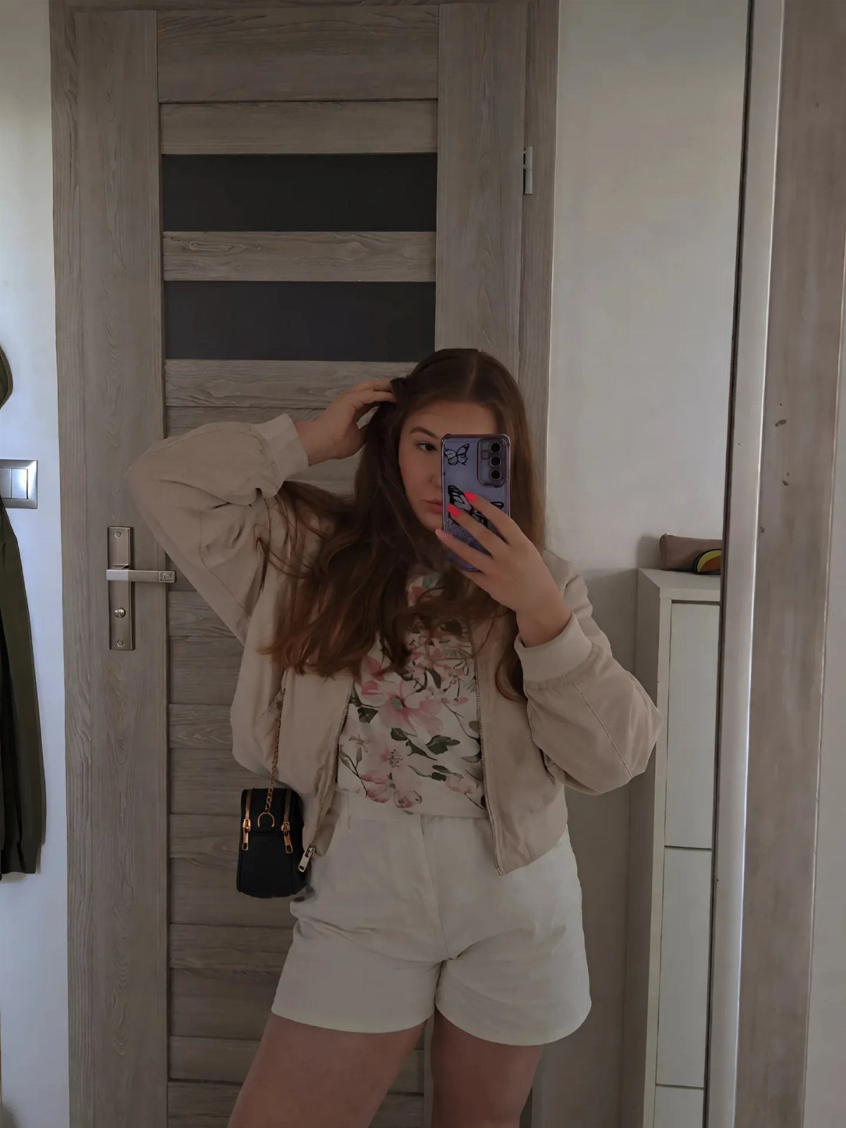 Young woman posing in a mirror wearing a floral top, white shorts, and a beige jacket.