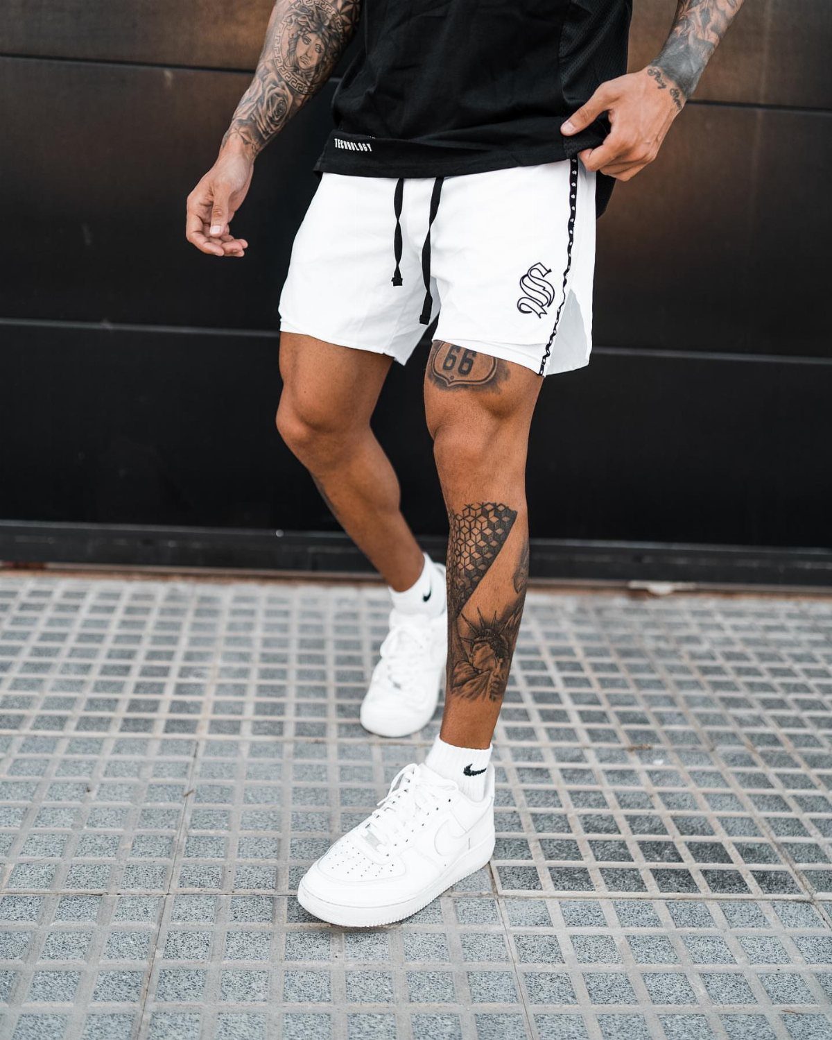 Man in white athletic shorts, black t-shirt, and white sneakers standing on a tiled ground.