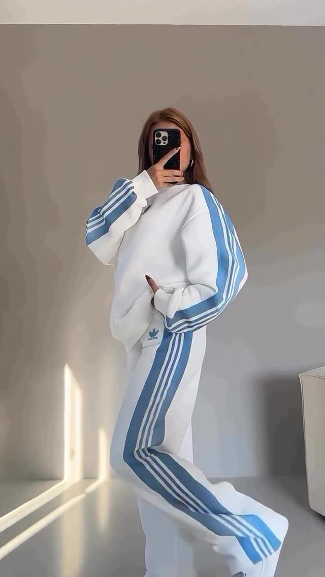 Woman in a white and blue striped tracksuit posing for a mirror selfie against a grey wall.