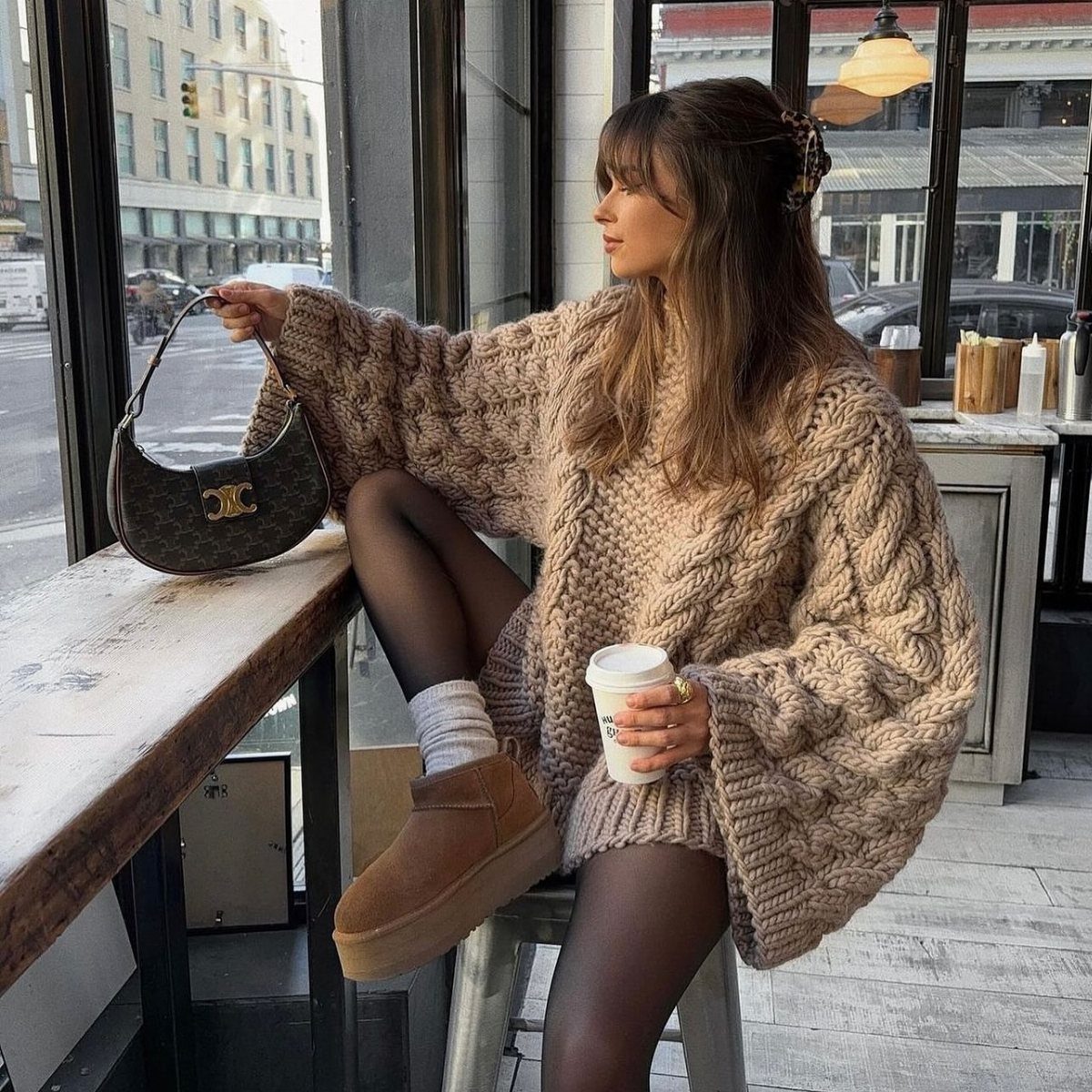 Woman wearing an oversized tan chunky knit sweater and black tights sitting by a window.