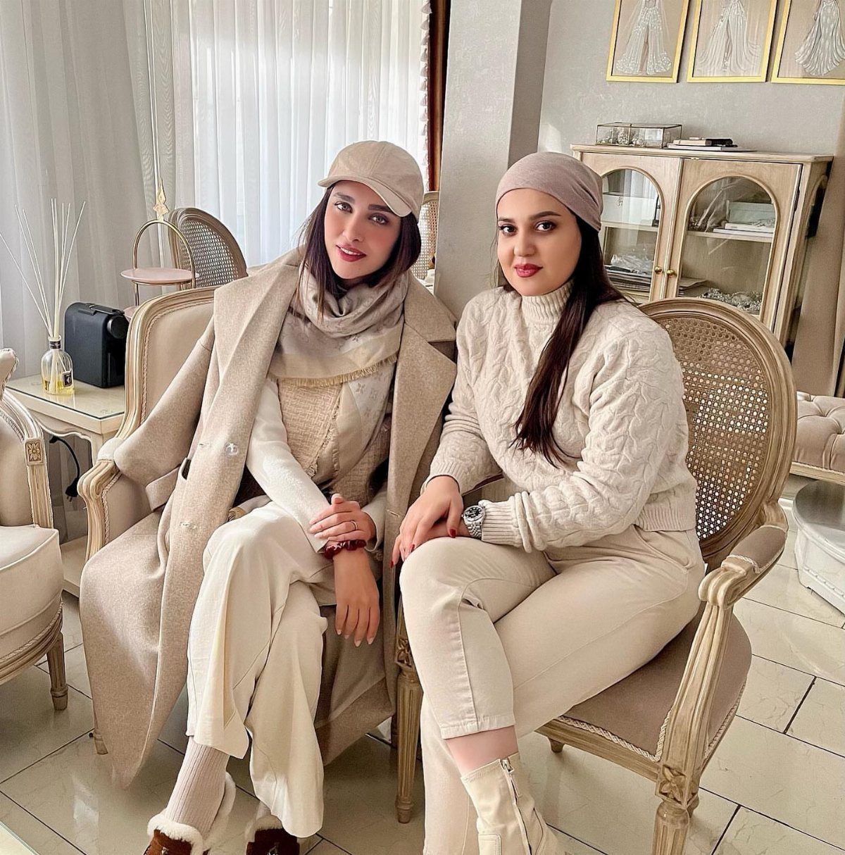 Two women posing in elegant, coordinated neutral-toned winter outfits while sitting in classic chairs.