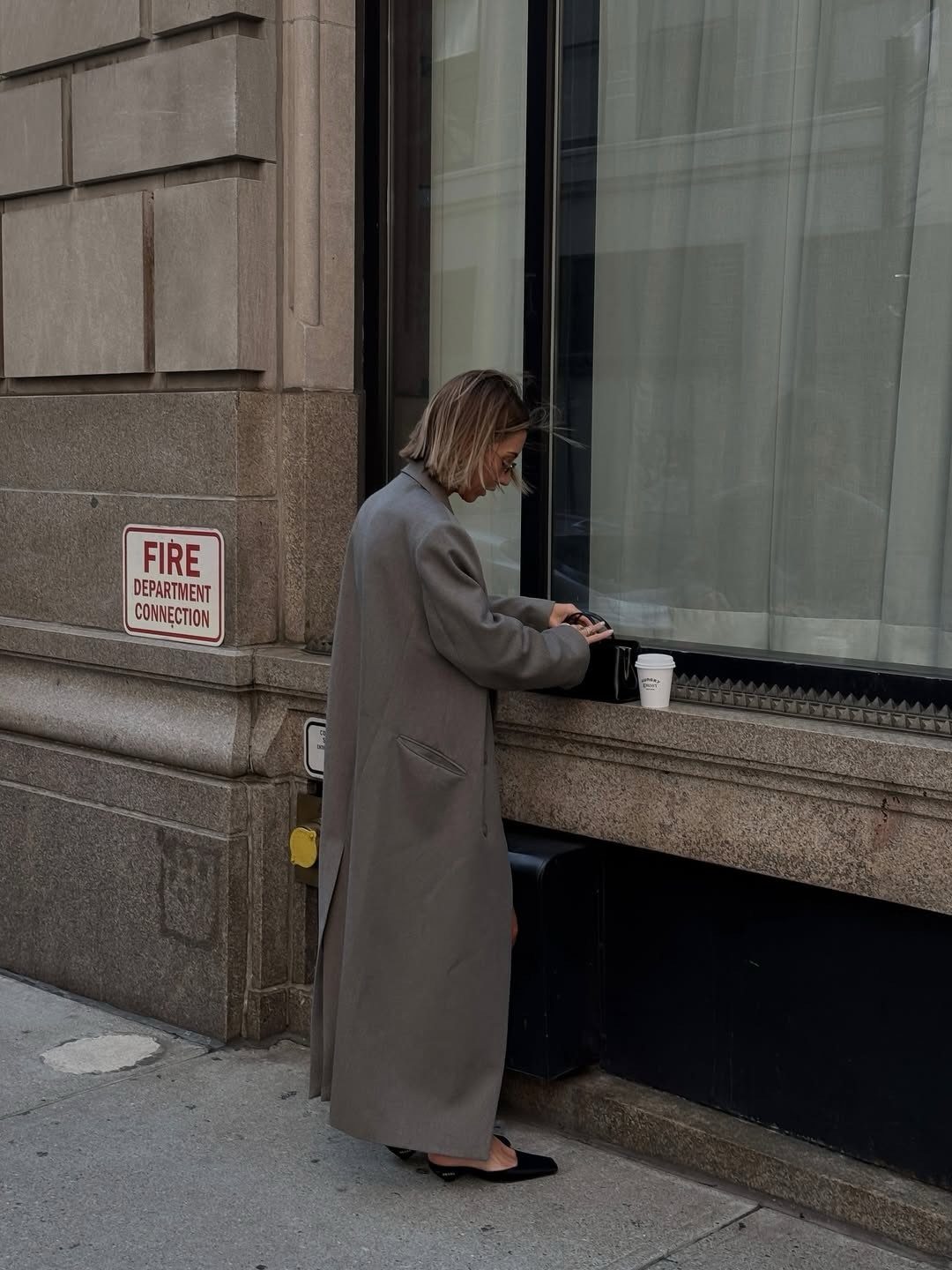 A woman in a long grey wool coat standing on a city sidewalk near a window.