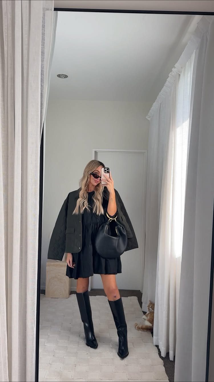 Woman in a black mini dress and boots wearing an olive green jacket draped over her shoulders.