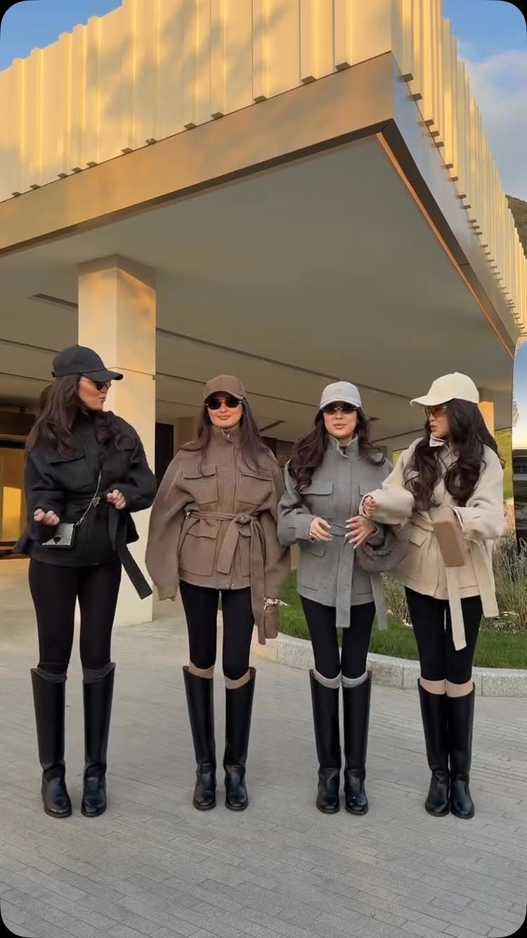 Four women standing together wearing neutral jackets, black leggings, and tall black riding boots.