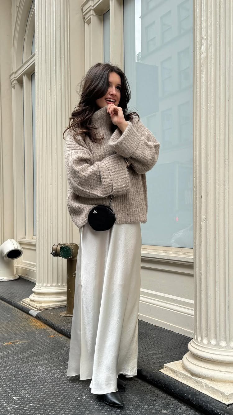 A smiling woman wearing a beige turtleneck sweater and a long white skirt standing outdoors.