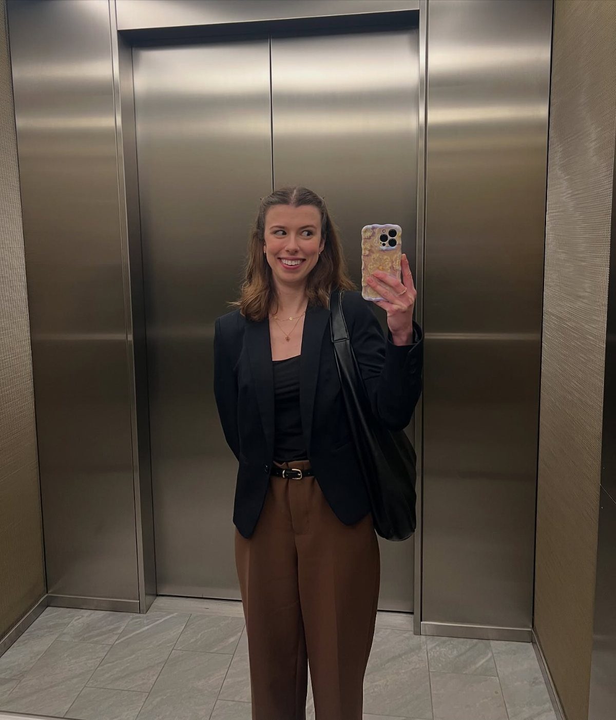 A woman smiling while taking a selfie in an elevator wearing a black blazer and brown trousers.