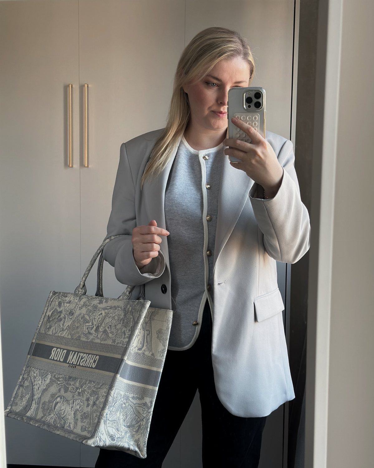 A woman posing in a mirror wearing a light blue blazer, white cardigan, and black pants while holding a large tote bag.