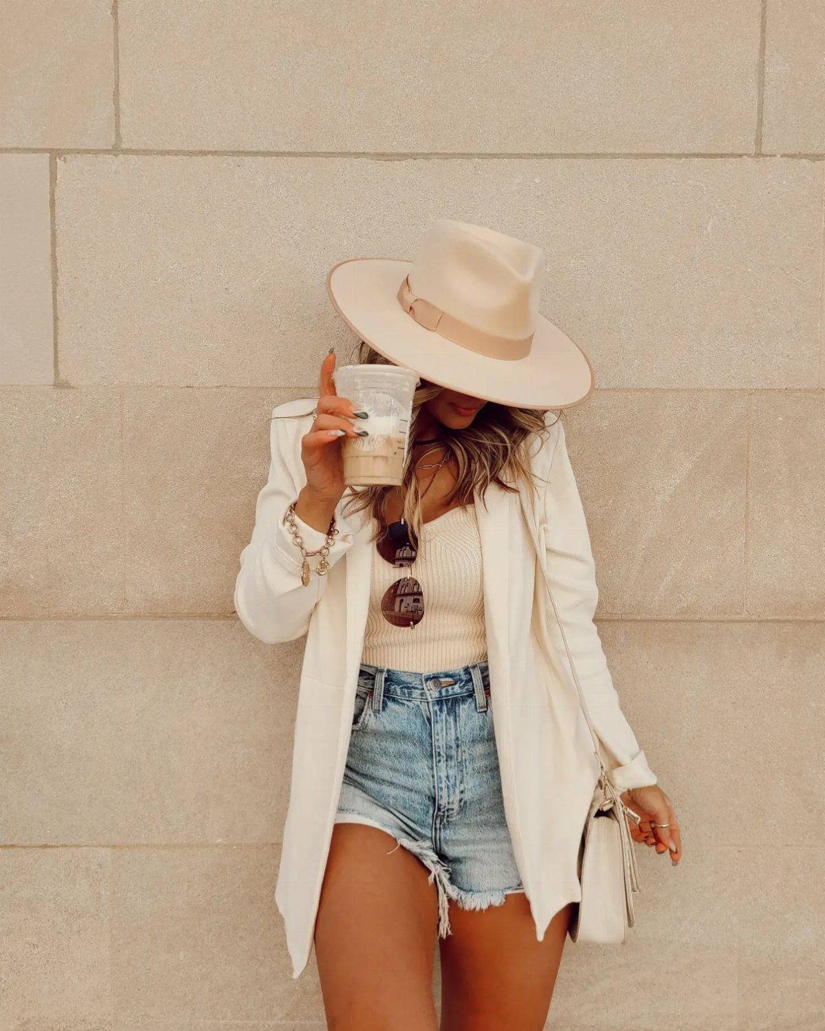 Woman in a white blazer and denim shorts holding a drink against a stone wall.