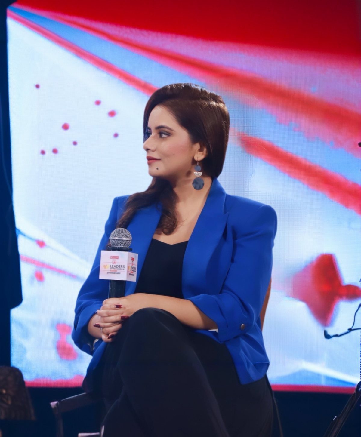 A woman wearing a bright blue blazer and black clothing sits holding a microphone.