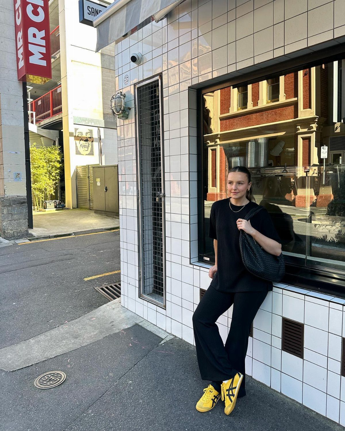 Woman in black athleisure clothing and bright yellow sneakers leaning against a white tiled wall.