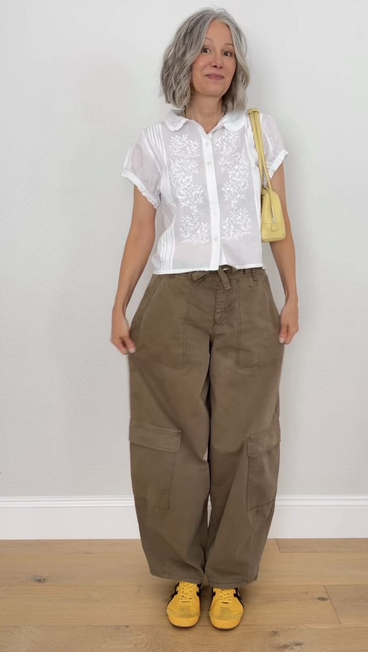 Woman wearing white shirt, olive cargo pants, and bright yellow sneakers standing against a white wall.