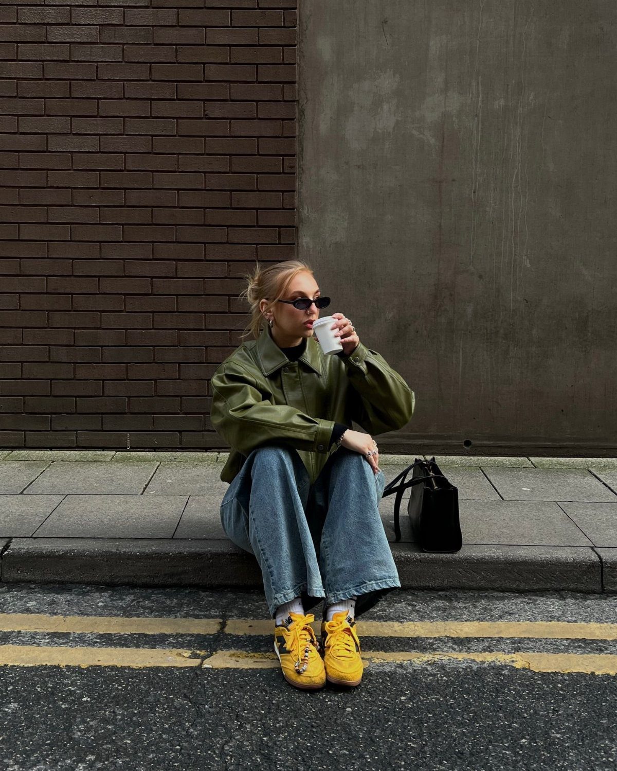 Person in an olive green jacket and blue jeans wearing bright yellow sneakers while sitting on a curb.