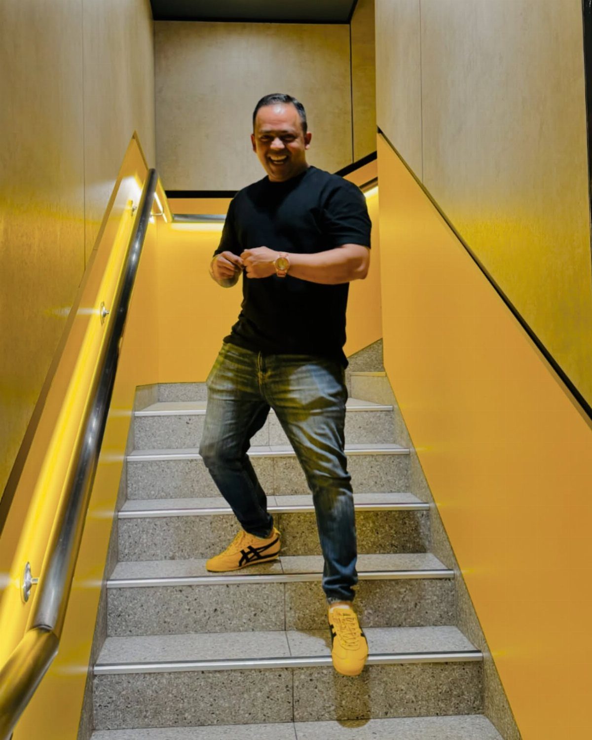 A man in a black t-shirt and blue jeans posing on yellow stairs wearing yellow sneakers.