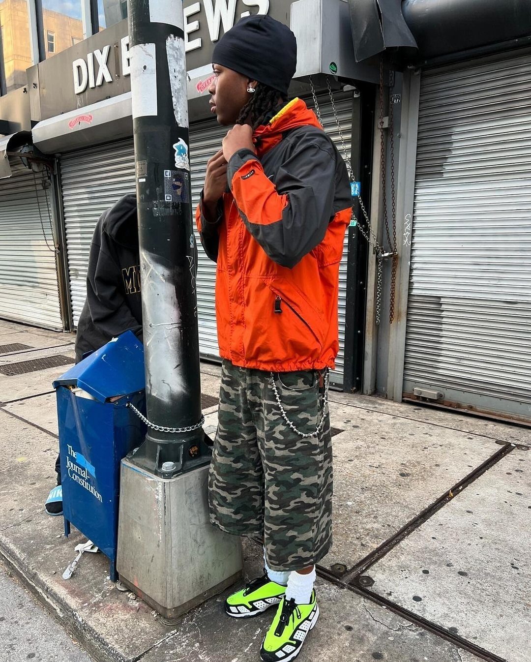 Person in orange jacket and camo shorts wearing bright neon yellow sneakers on a city street.