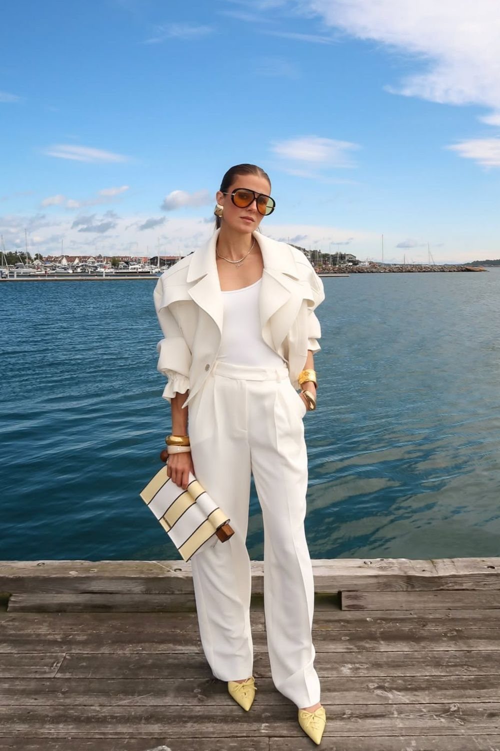 Woman in an all-white outfit and yellow shoes standing on a wooden pier by the ocean.